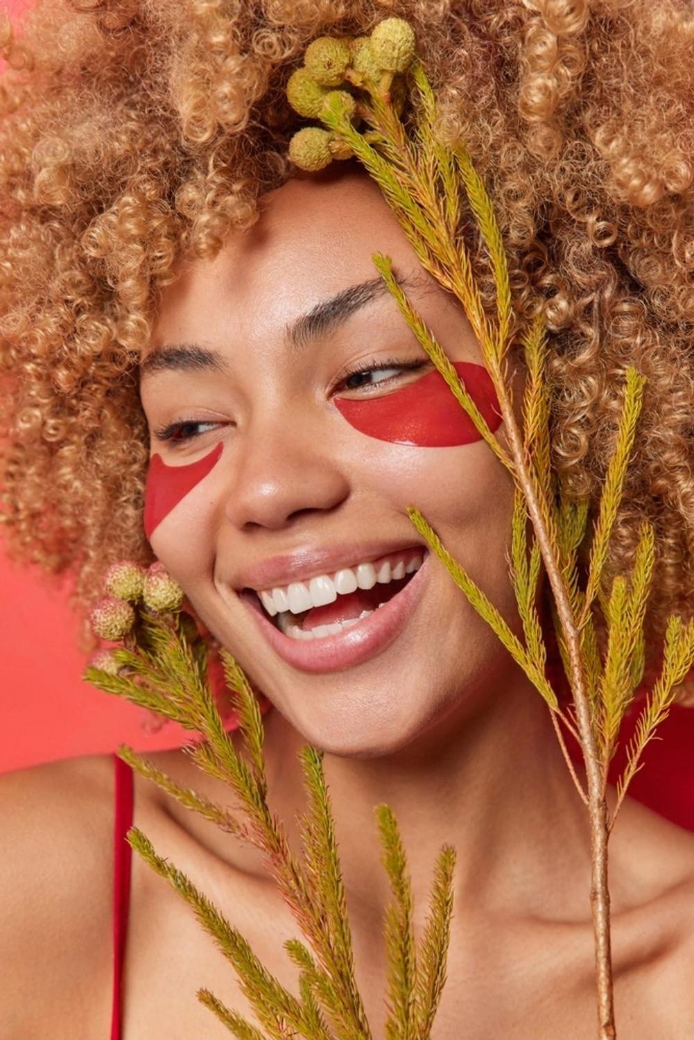 Woman with red under-eye patches smiling next to green botanical branches