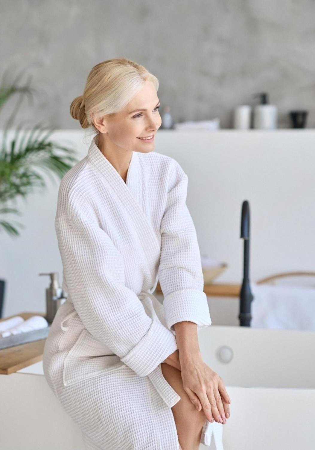 Woman in a white robe relaxing beside a bathtub in a bright spa setting